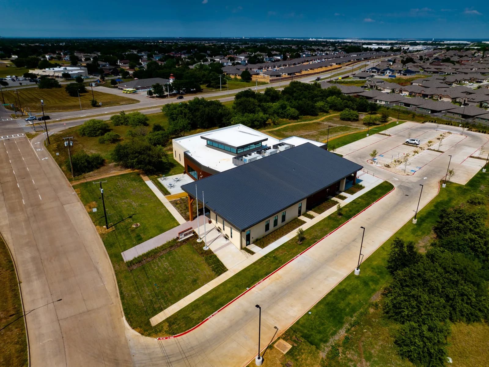 Aerial view of a modern low-rise building with a dark roof, surrounded by a parking lot and nearby houses