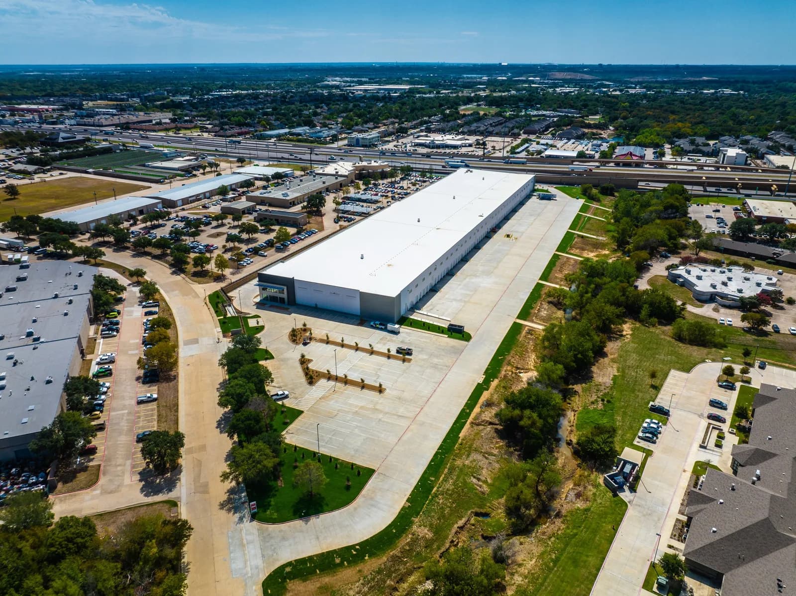 Aerial view of a large white-roofed warehouse with parking lots and an adjacent highway