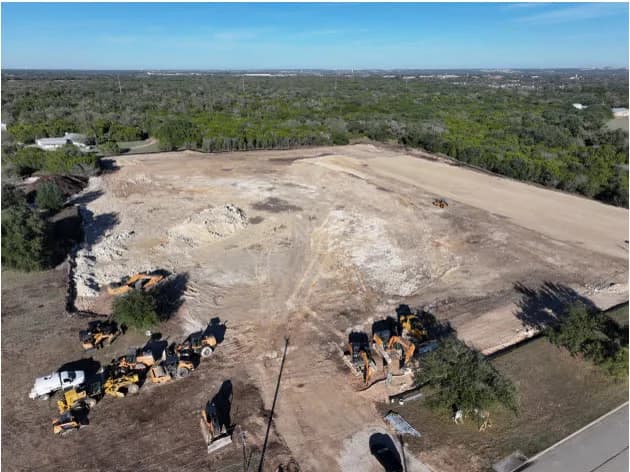 Aerial view of a construction site with dirt piles, excavators and dump trucks, surrounded by trees