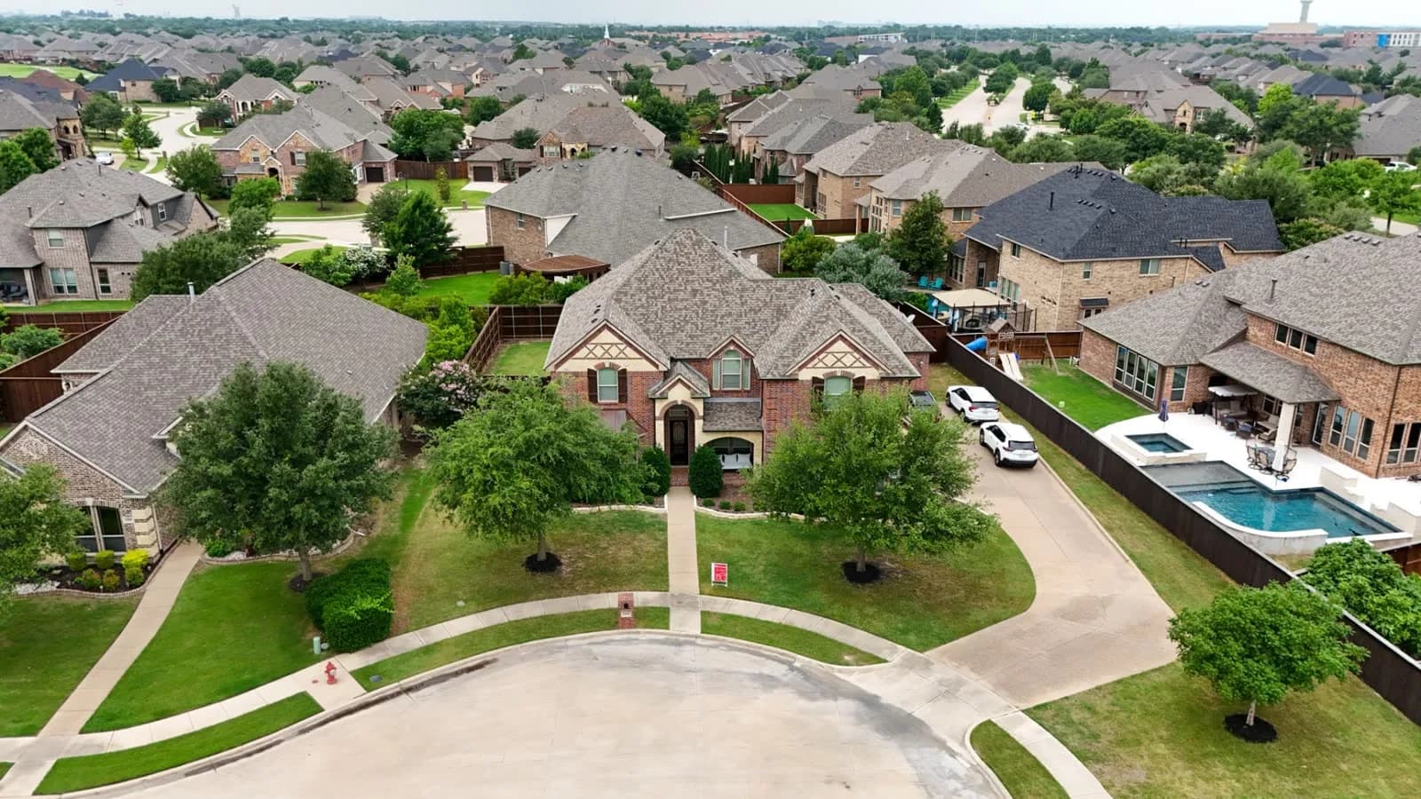 Drone view of a suburban neighborhood with brick homes, a central two-story house, green lawns, and a pool