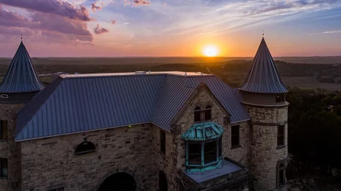 Sunset over a stone mansion with slate roof and two turret towers, turquoise glass bay