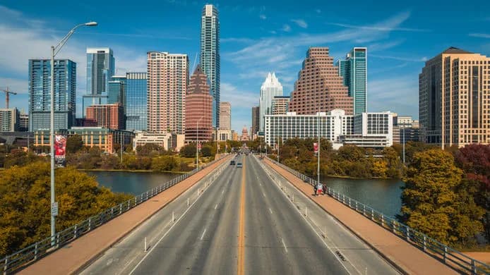 Bridge leads to a city skyline with tall buildings across a river, flanked by trees
