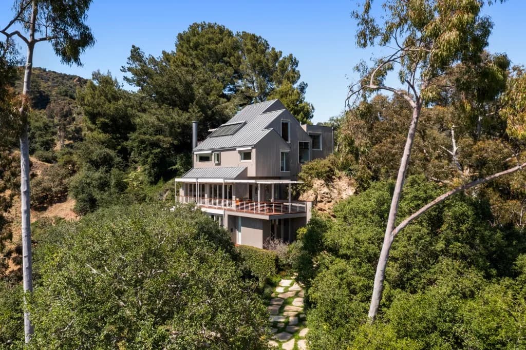 Modern multi-level house with wraparound deck and stone path among trees on a hillside