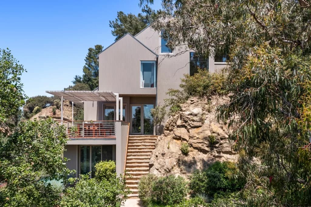 Modern beige house built into a rocky hillside with a wooden deck, pergola and stairs