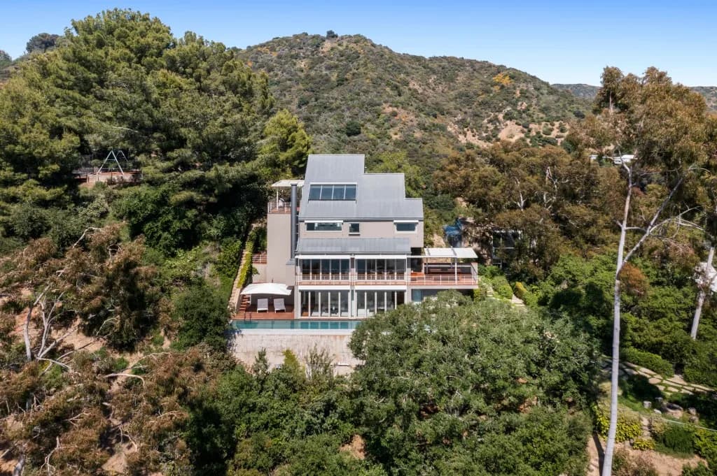 Modern multi-level house with large windows and balconies among trees on a hillside, with a pool in front