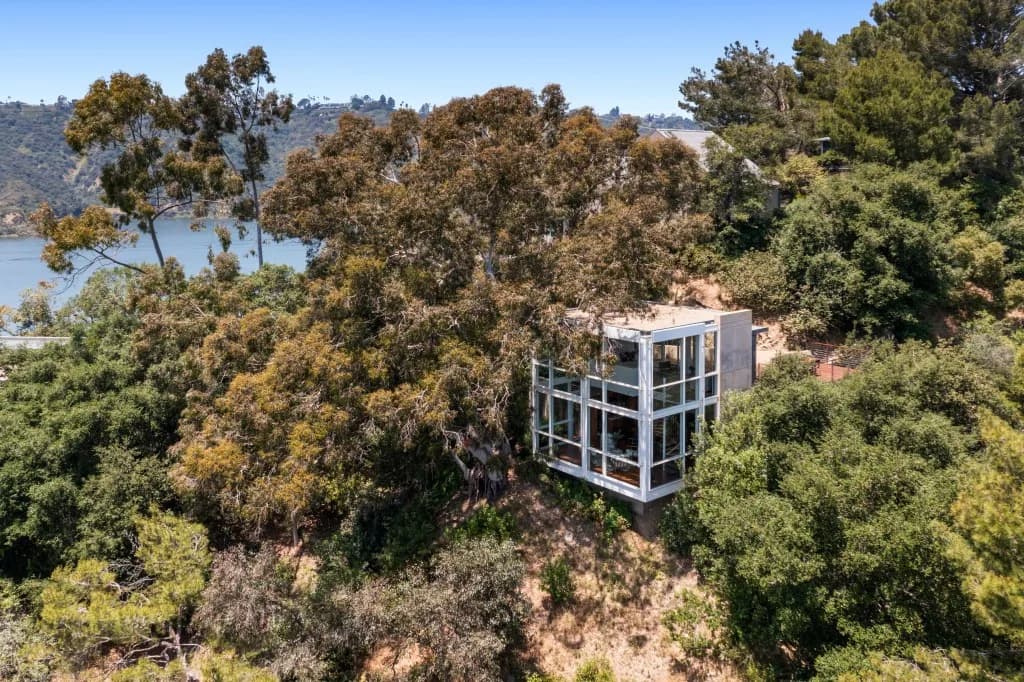 Glass-walled cubic house on a hillside amid trees, with water and hills in the background