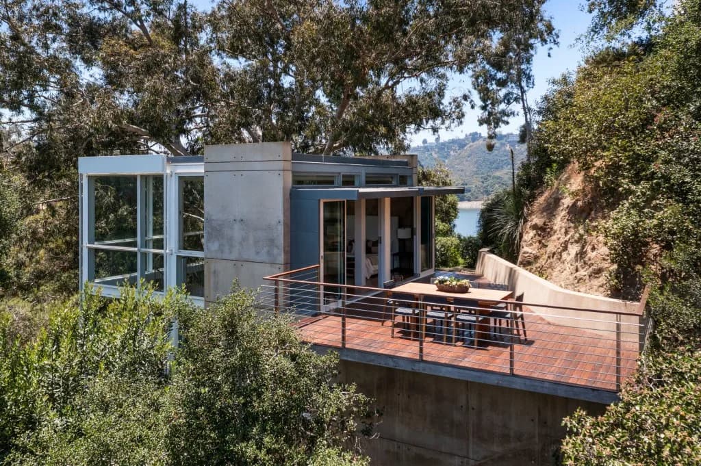 Modern glass and concrete cabin on a hillside with a wooden deck, metal railing, and outdoor dining table