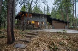Two-story wooden cabin in a forest with a garage, stone steps, and lit windows