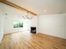 Bright living room with white walls, hardwood floor, fireplace, chandelier, ceiling beam, and large window