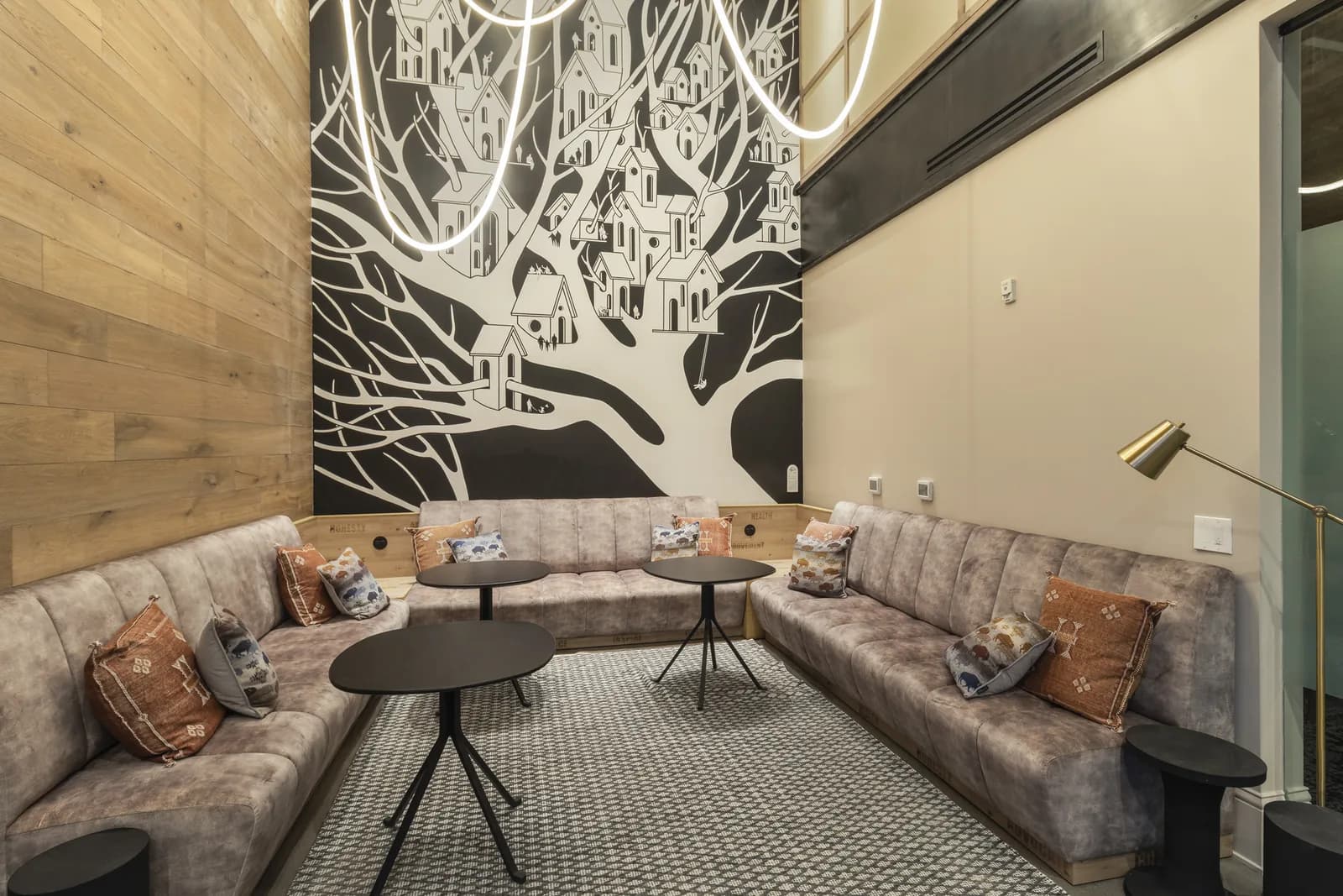 Cozy lounge with gray sectional sofa, round black tables, patterned rug, and black-and-white tree mural