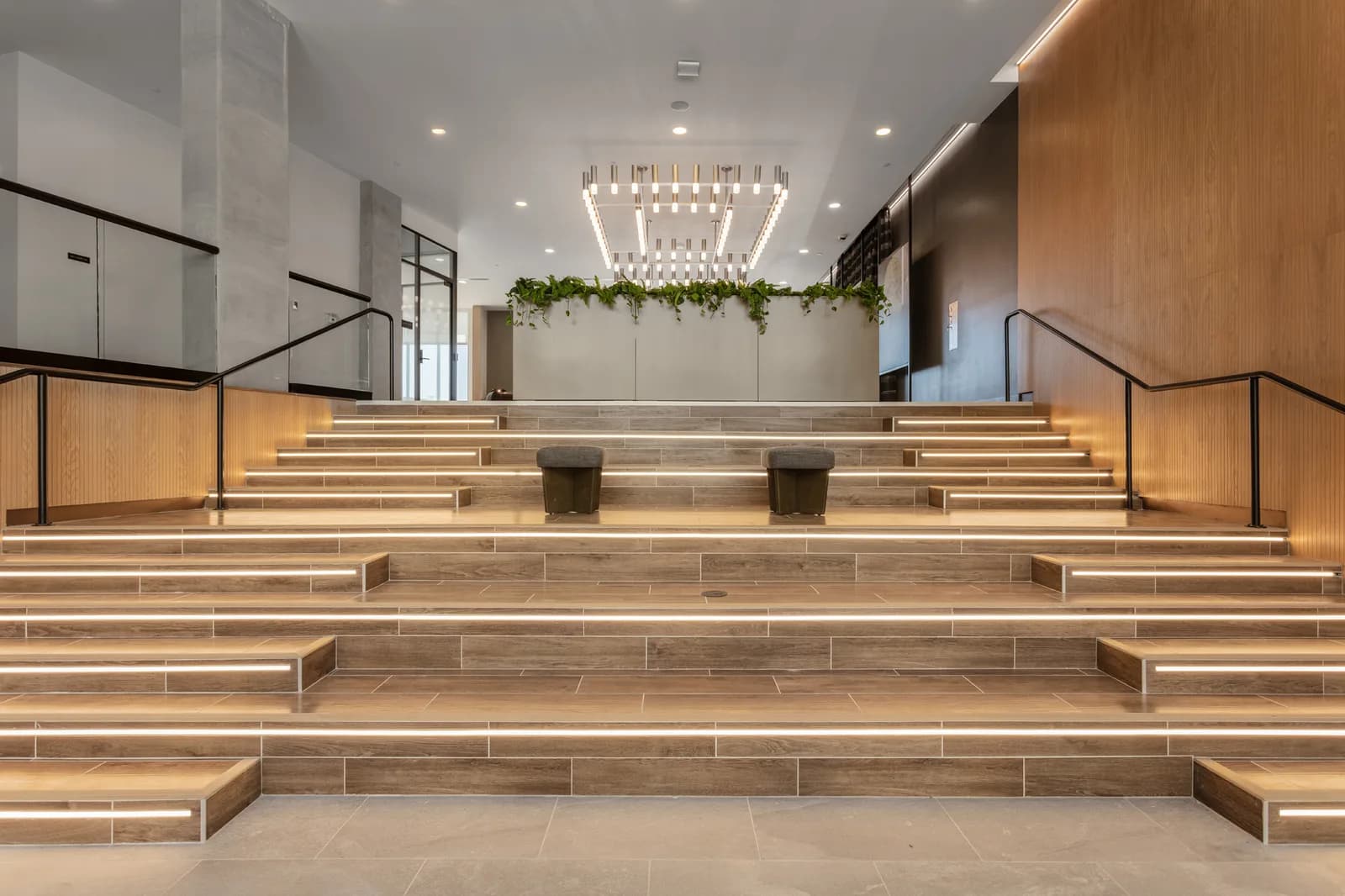 Interior lobby with LED-lit steps, wood walls, a planter with greenery, two ottomans, and a modern chandelier