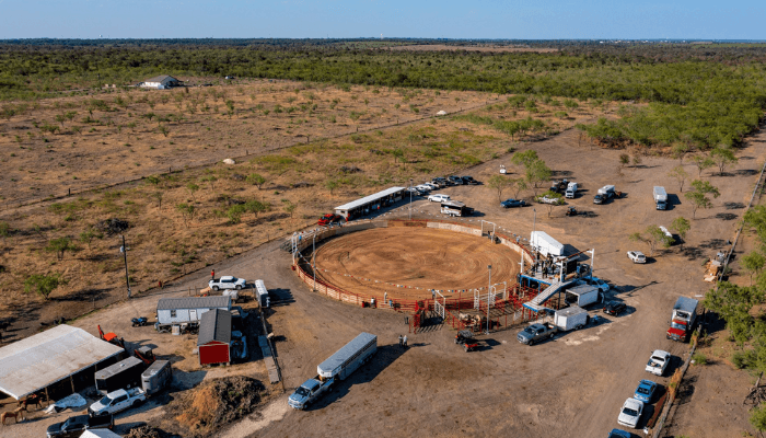 Aerial view of a circular dirt arena fenced in, with trucks, trailers and tents in a sparse desert landscape