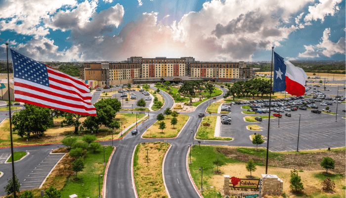 Aerial view of a large campus with curved roads, green lawns, and two flags (US and Texas) in the foreground