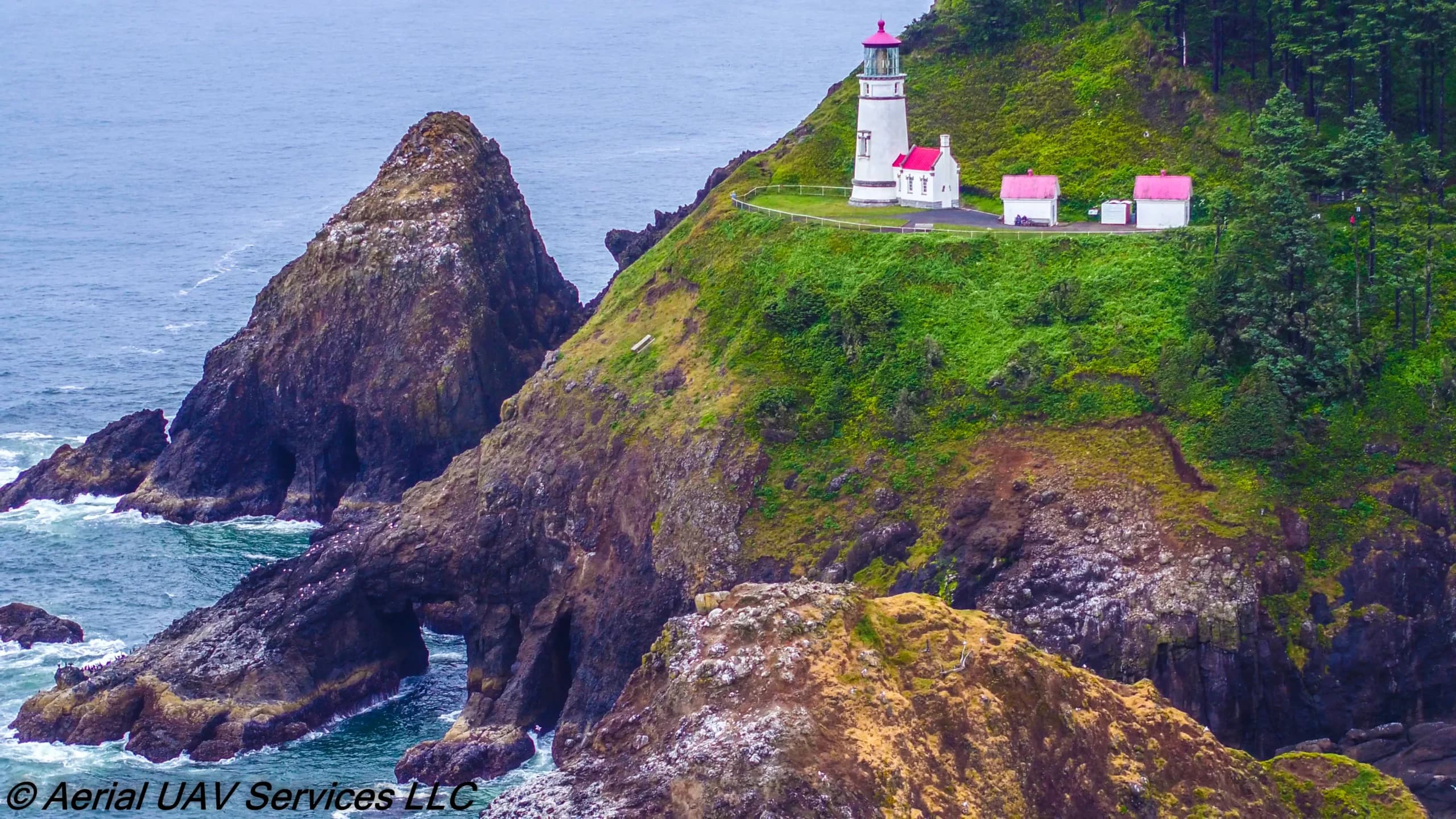 Lighthouse on a green cliff with white buildings and pink roofs, overlooking the rocky shoreline