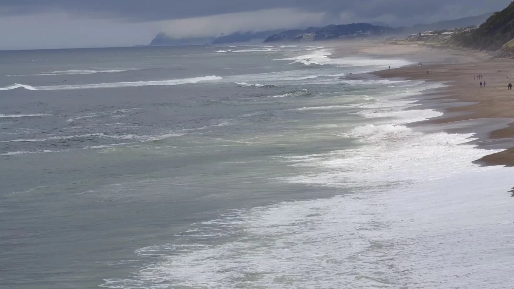 Coastal scene of a sandy beach with waves, distant hills, and a cloudy sky with people along the shore