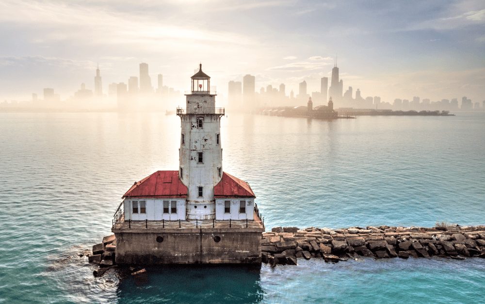 White lighthouse with a red roof on a rocky jetty, hazy city skyline across calm water