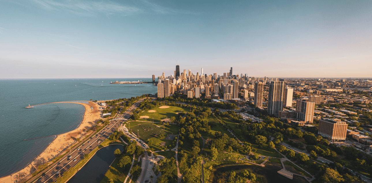 An aerial view of a lakefront city skyline with a green park and a sandy beach along the shore