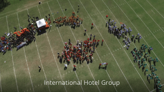 Aerial view of colored groups of people lined up on a grass field with white lines and a white tent