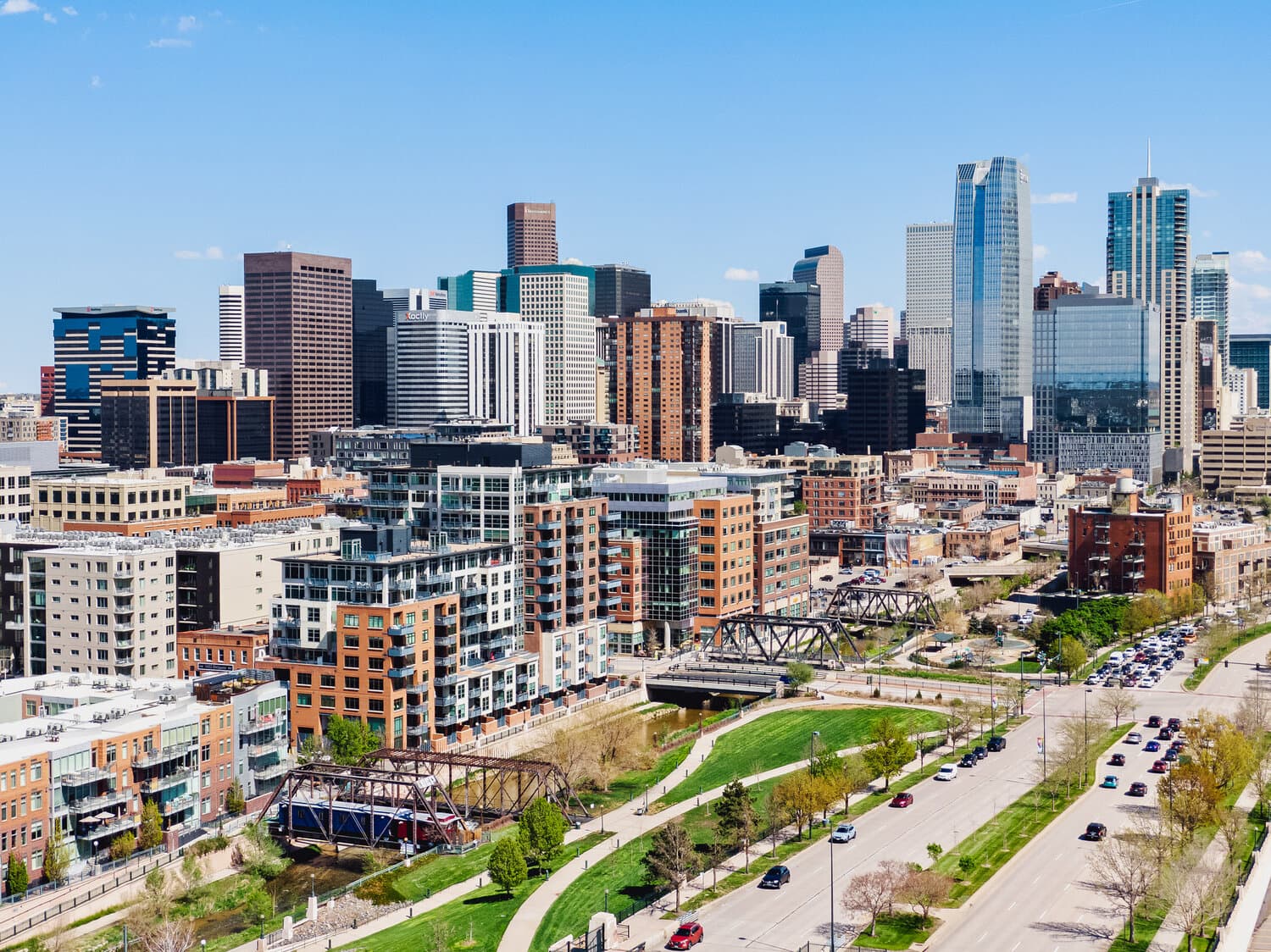 City skyline with tall skyscrapers, mid-rise buildings, a riverfront park, and a busy road in the foreground