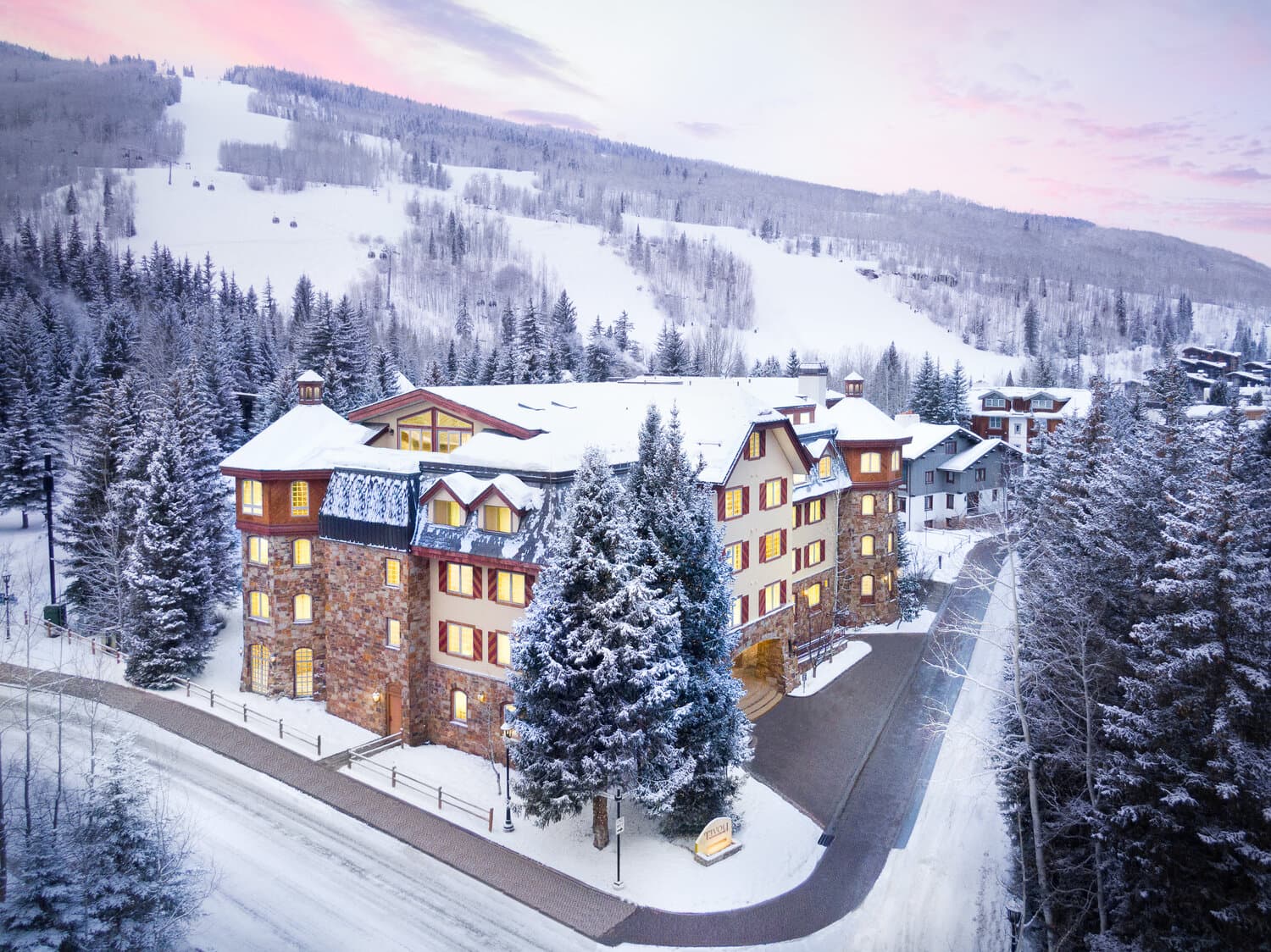 Snowy alpine hotel complex with stone and wood facade, lit windows, surrounded by snow-covered evergreens