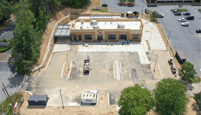 An aerial view of a single-story beige commercial building with a large concrete lot and surrounding trees
