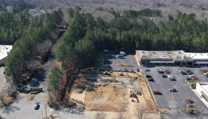 Aerial view of a construction site with an excavator and dirt pit beside a parking lot and trees