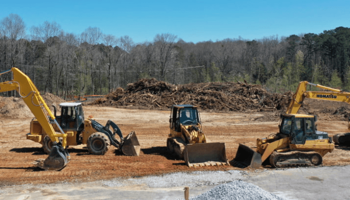 Three yellow construction excavators and loaders on a dirt site with a wood pile and trees in the background