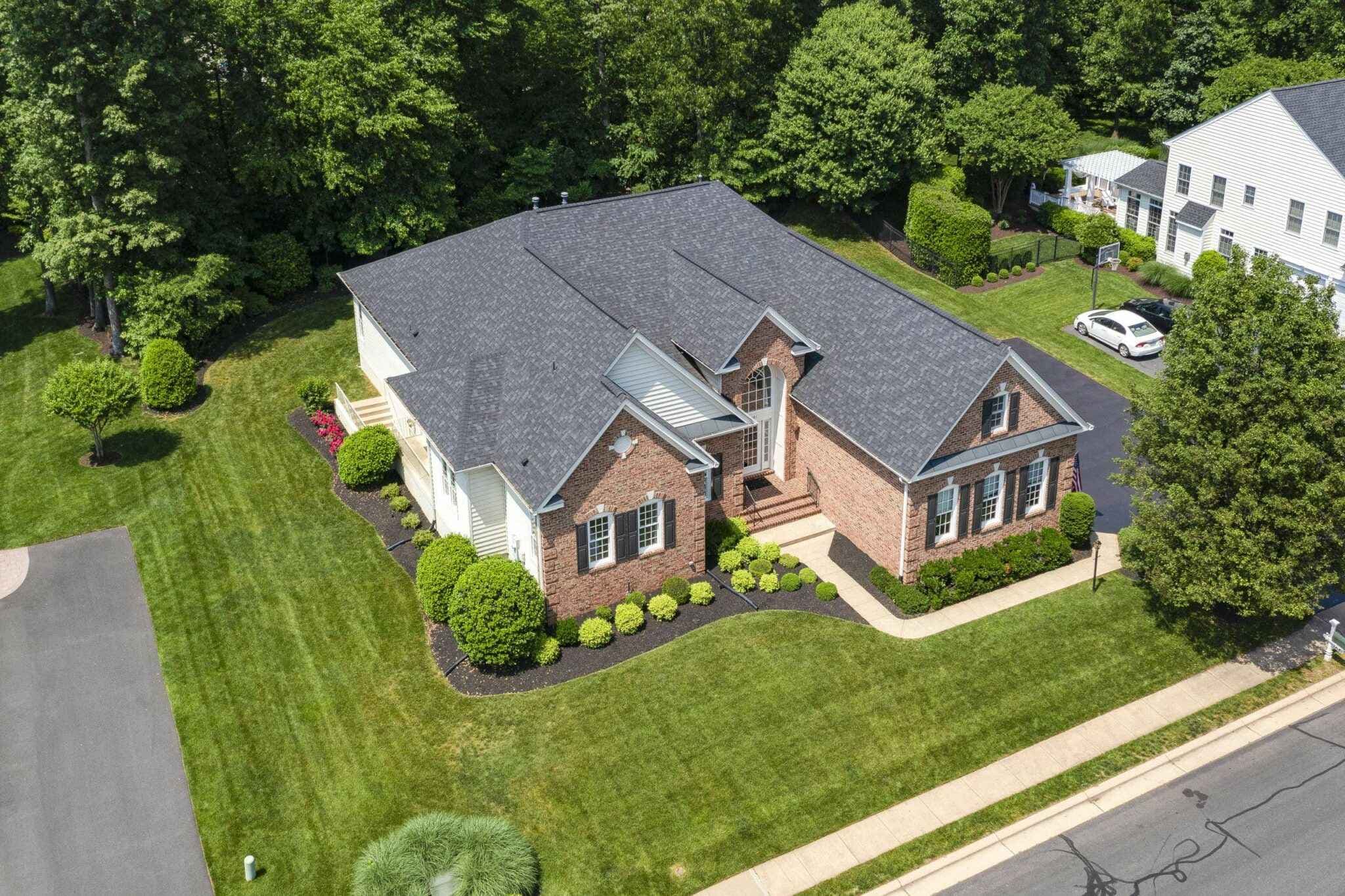 Aerial view of a brick house with a gray shingle roof, manicured lawn, and adjacent white house