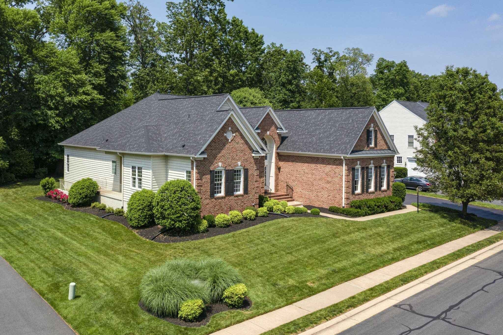 Brick and siding house with a gray roof, arched entry, manicured lawn, and surrounding trees