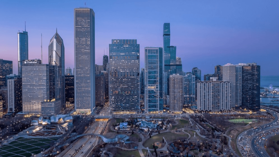 City skyline with tall glass skyscrapers, river on the right, and a park with winding paths in the foreground