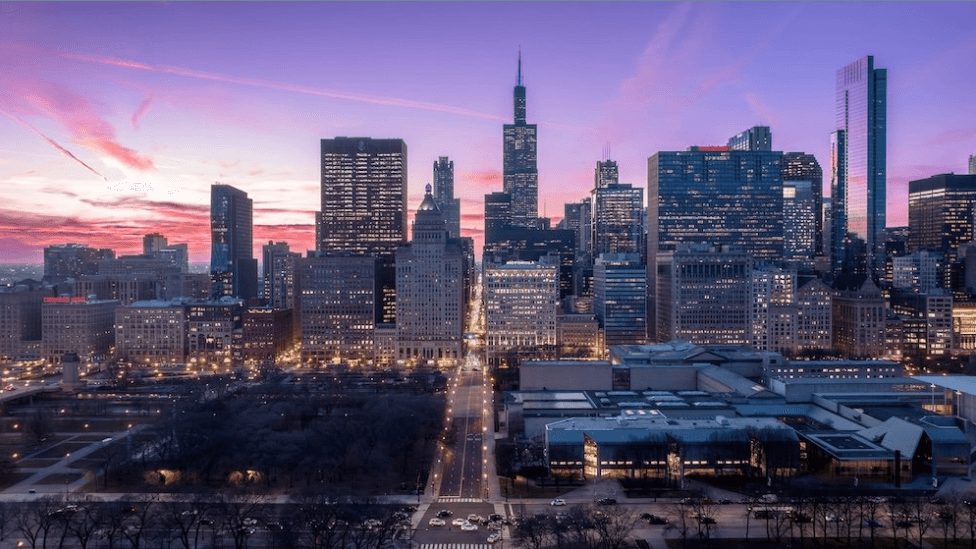 City skyline at dusk with tall buildings, pink and purple sky, lit streets, and a foreground park
