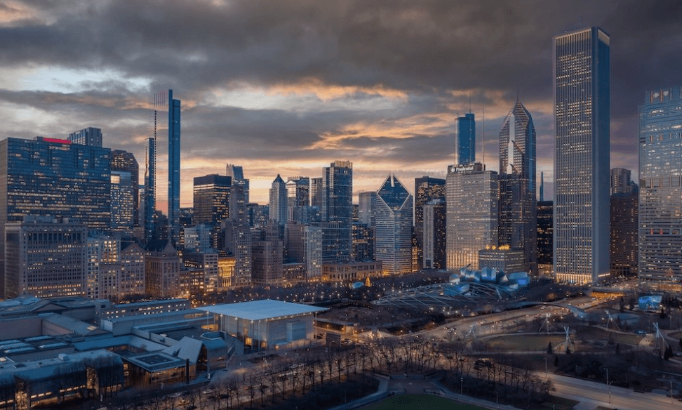 Dusk city skyline with tall glass skyscrapers, orange sunset sky, and a park in the foreground