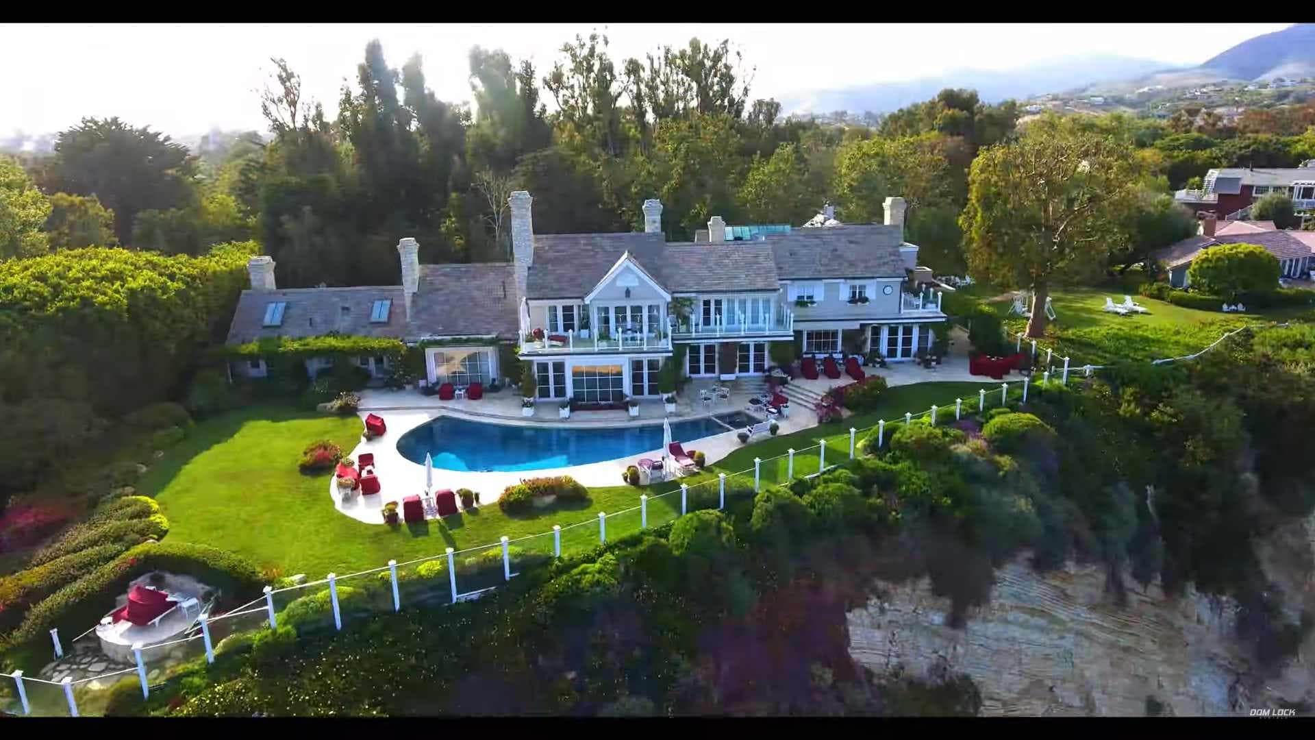 Aerial view of a large house with chimneys, a curved pool, manicured lawn, and red seating