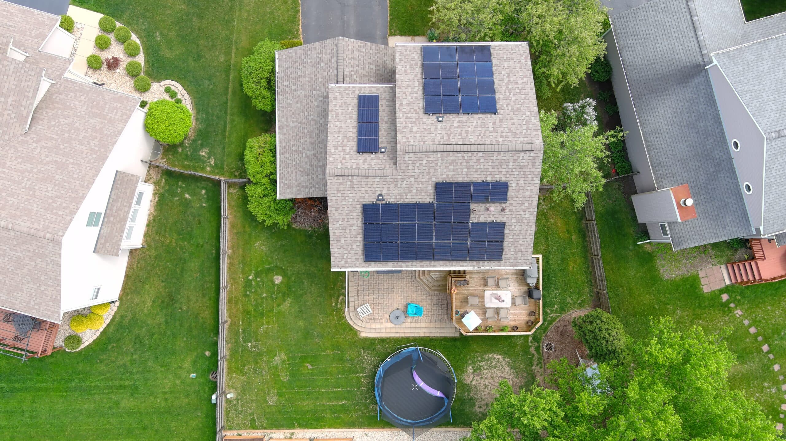 Aerial view of a suburban home with numerous solar panels on the roof, surrounded by green lawns and trees