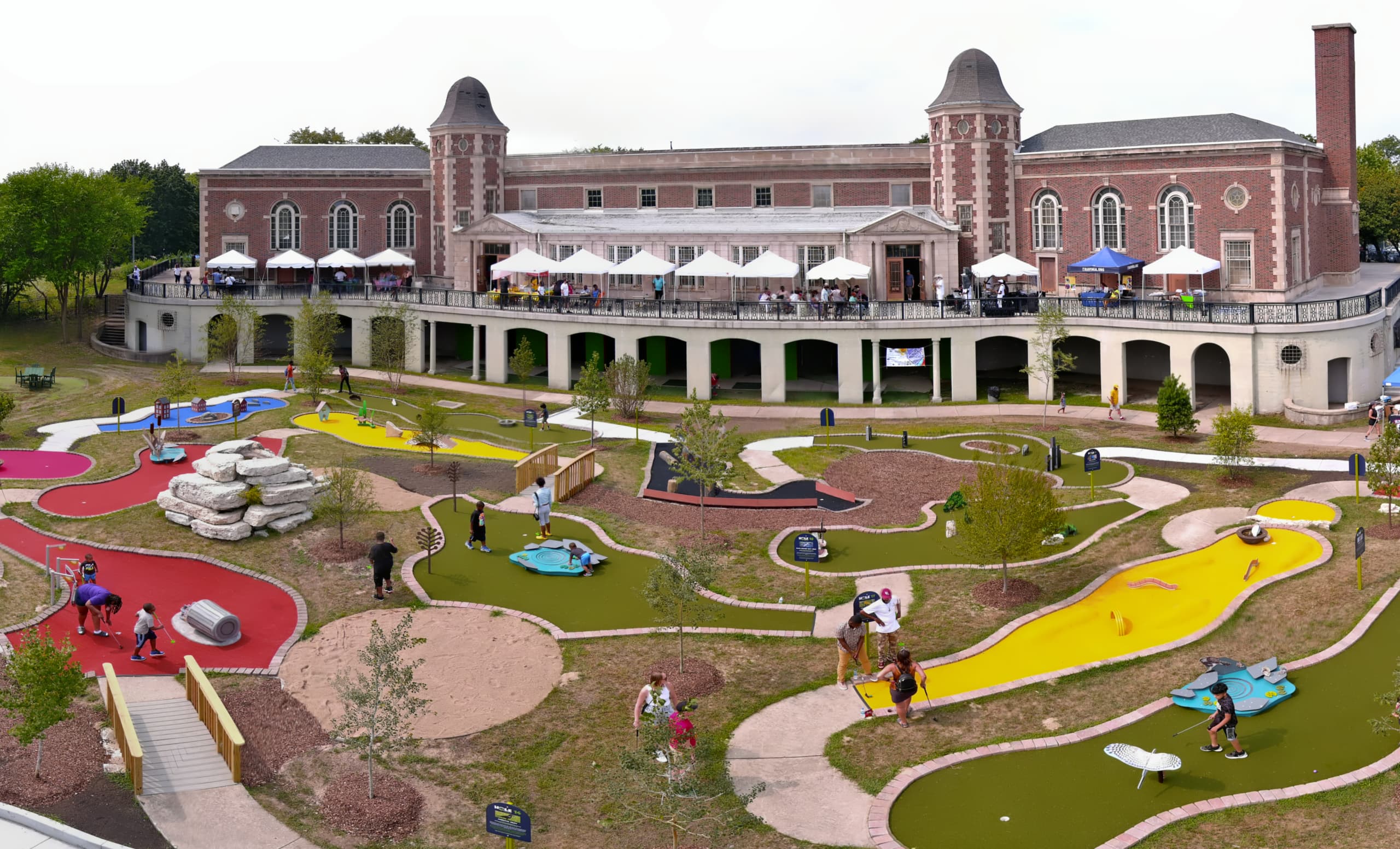 Brick building with arched windows and towers, terrace with white umbrellas, and a colorful mini golf course
