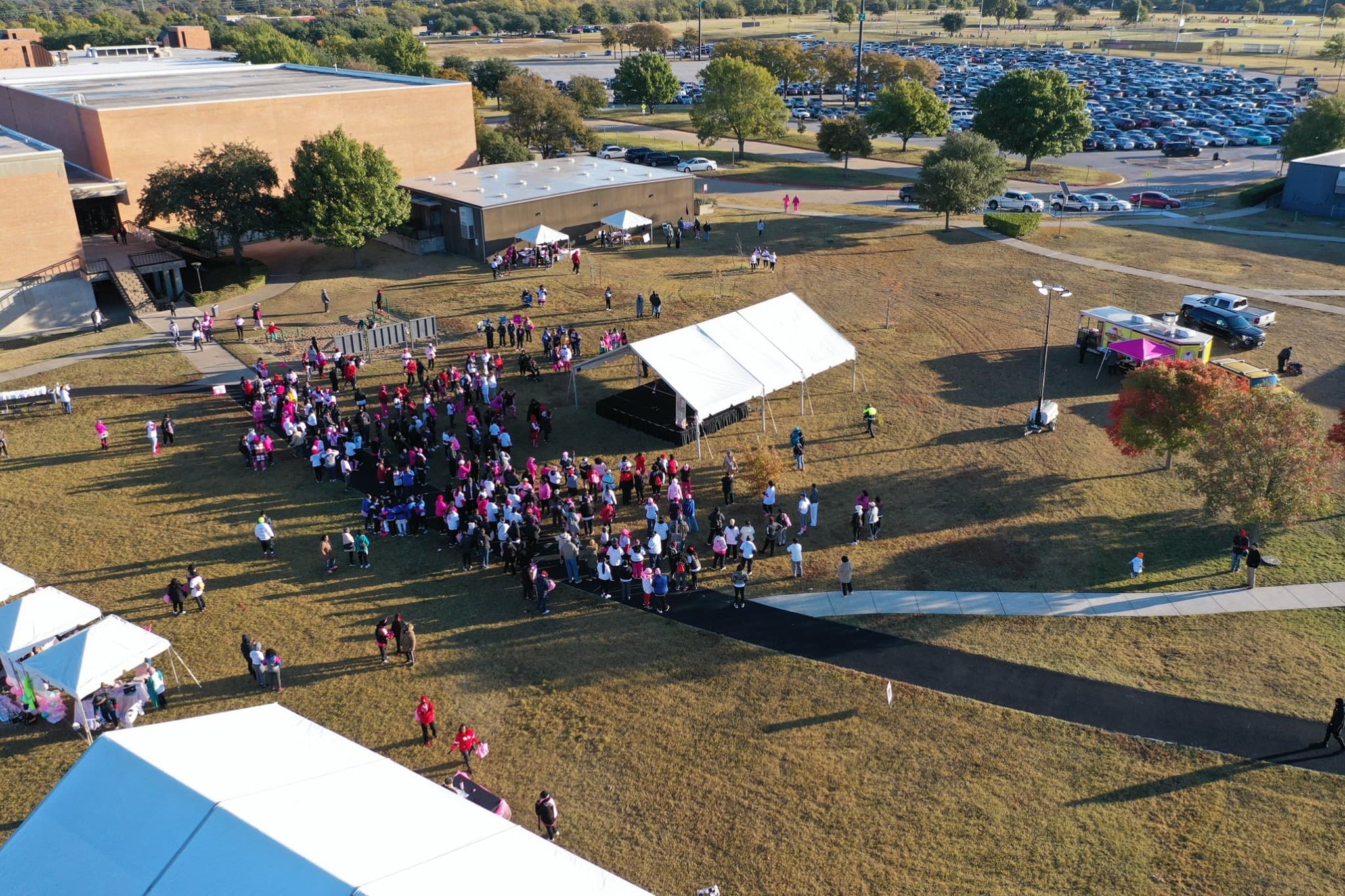 Aerial view of an outdoor event at a large brick building, with a crowd, tents, and a crowded parking lot
