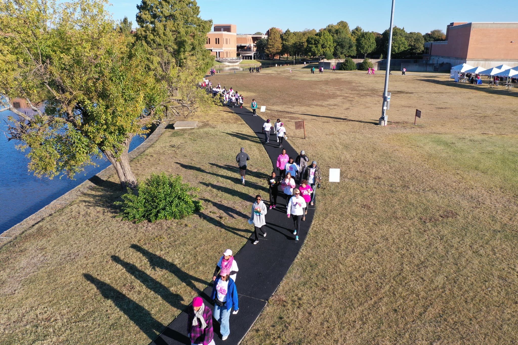Aerial view of a crowd walking along a curved black path through a park by a river, with trees and tents