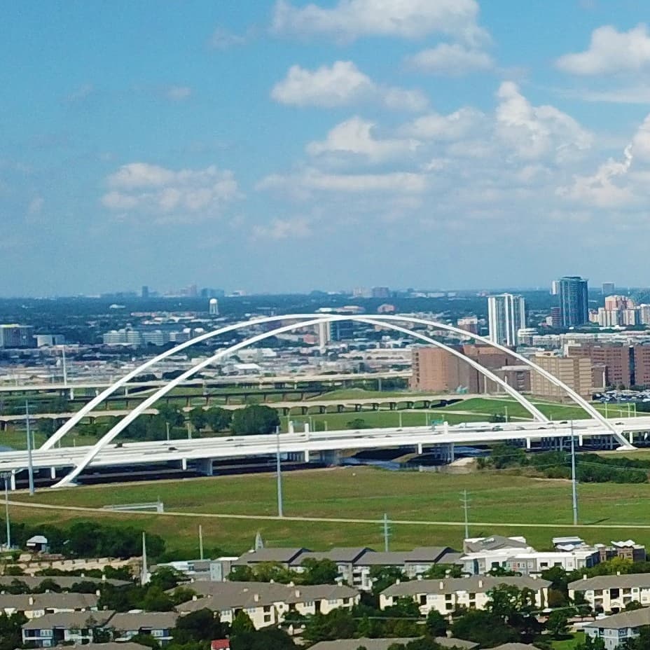 White arch bridge spans a highway over green fields with a city skyline in the distance