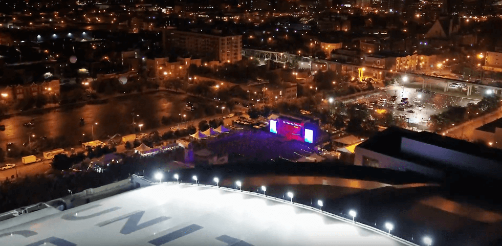 Night aerial view of a city with tents, stage and screens, and a large curved white surface in the foreground