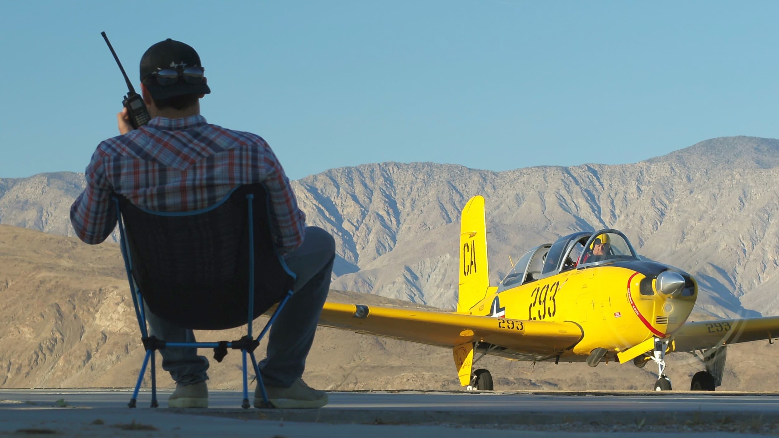Man in plaid shirt on a folding chair with a radio, next to a yellow prop plane, mountains in the distance