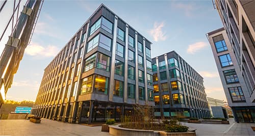 Row of modern glass office buildings around a paved plaza with planters at sunset