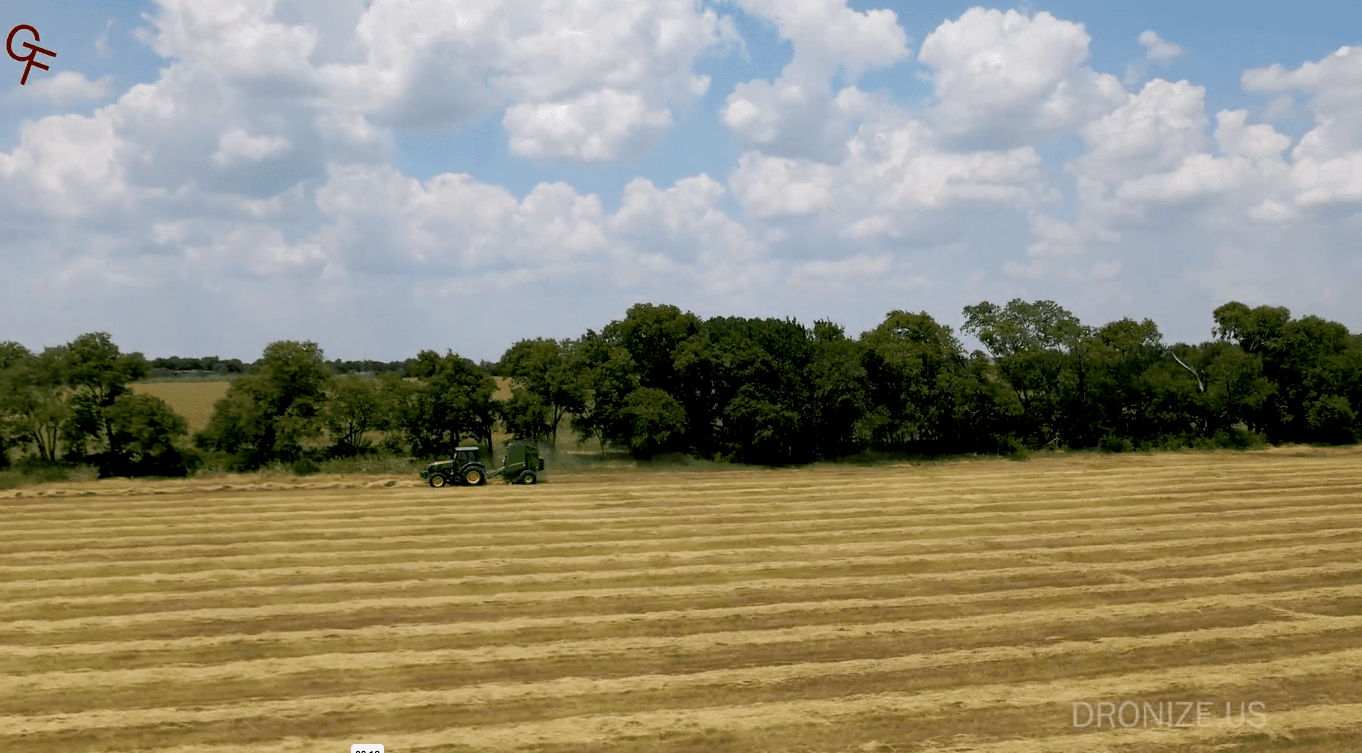 Red logo top-left, green tractor with baler in striped hay field and cloudy sky; DRONIZE.US watermark