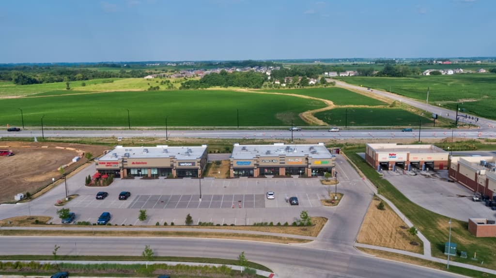 Aerial view of a strip mall with storefronts and a large parking lot, adjacent road and green fields