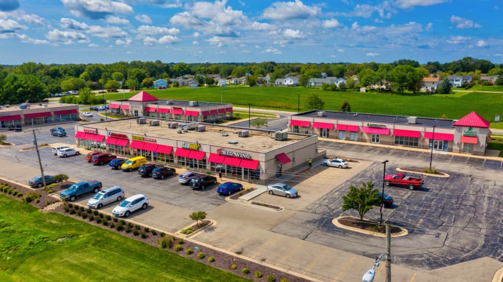 Aerial view of a strip mall with red awnings, many parked cars, and surrounding green fields