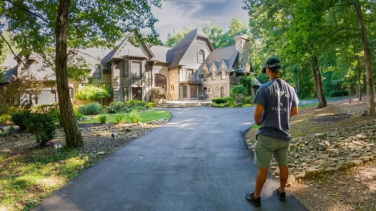 Drone operator on a curved driveway toward a large stone mansion surrounded by trees