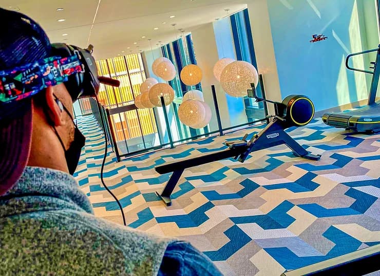Person with headband and mask watches a rowing machine in a bright room with globe lights, a drone in view