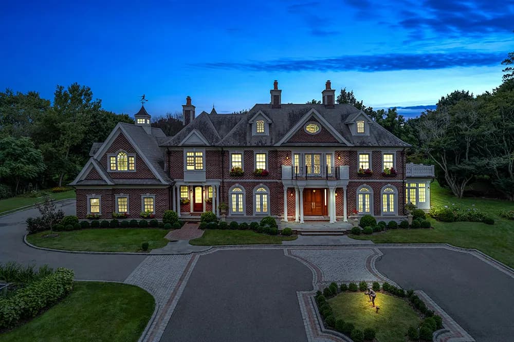 Aerial view of a large brick mansion at dusk with circular driveway and lit windows