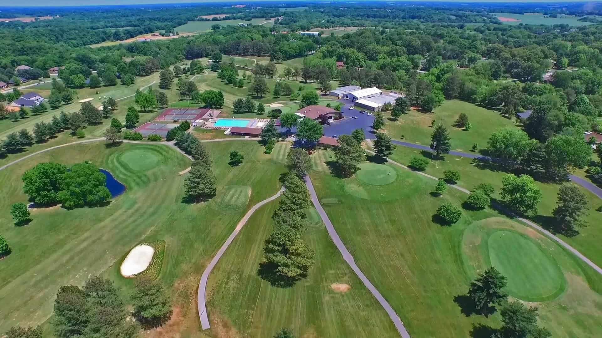 Aerial view of a golf course with greens, fairways, trees, a clubhouse, pool and tennis courts
