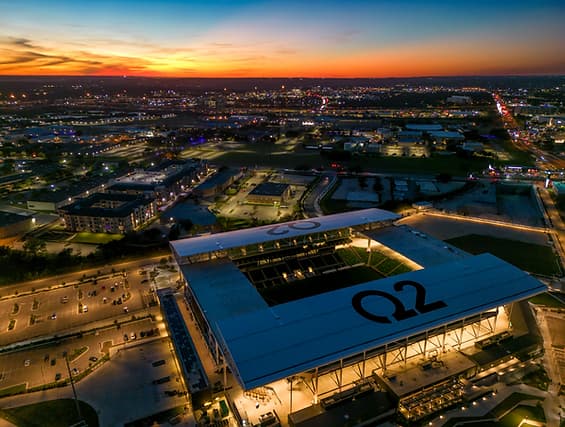 Aerial dusk view of a city with a large roofed arena bearing the Q2 logo and surrounding streets