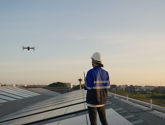 Person in a hard hat and blue reflective jacket stands on a solar roof, drone flying nearby